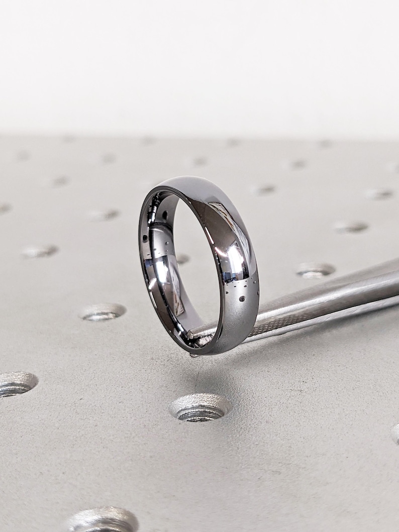 May include: A polished, silver-colored ring with a smooth, rounded design. The ring is held by a metal tool, showcasing its reflective surface. The ring's simple design suggests it could be a wedding band or a fashion accessory.