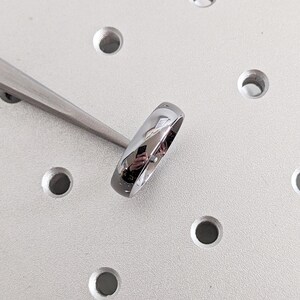 May include: A shiny, silver ring with a smooth, polished surface. The ring is held by a pair of silver tweezers against a light gray, industrial-looking background with several circular indentations. The ring reflects the surrounding environment.