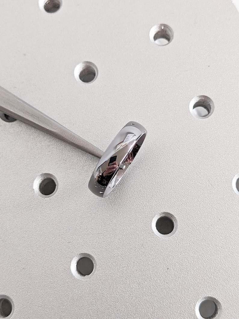 May include: A shiny, silver ring with a smooth, polished surface. The ring is held by a pair of silver tweezers against a light gray, industrial-looking background with several circular indentations. The ring reflects the surrounding environment.