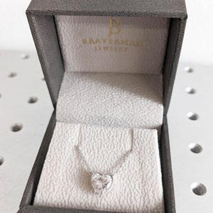 Puede incluir: Un collar de plata con un colgante en forma de coraz&oacute;n con un gran diamante transparente en una caja de joyer&iacute;a gris con el texto "BRAVERMAN JEWELLERY" en el interior de la tapa.