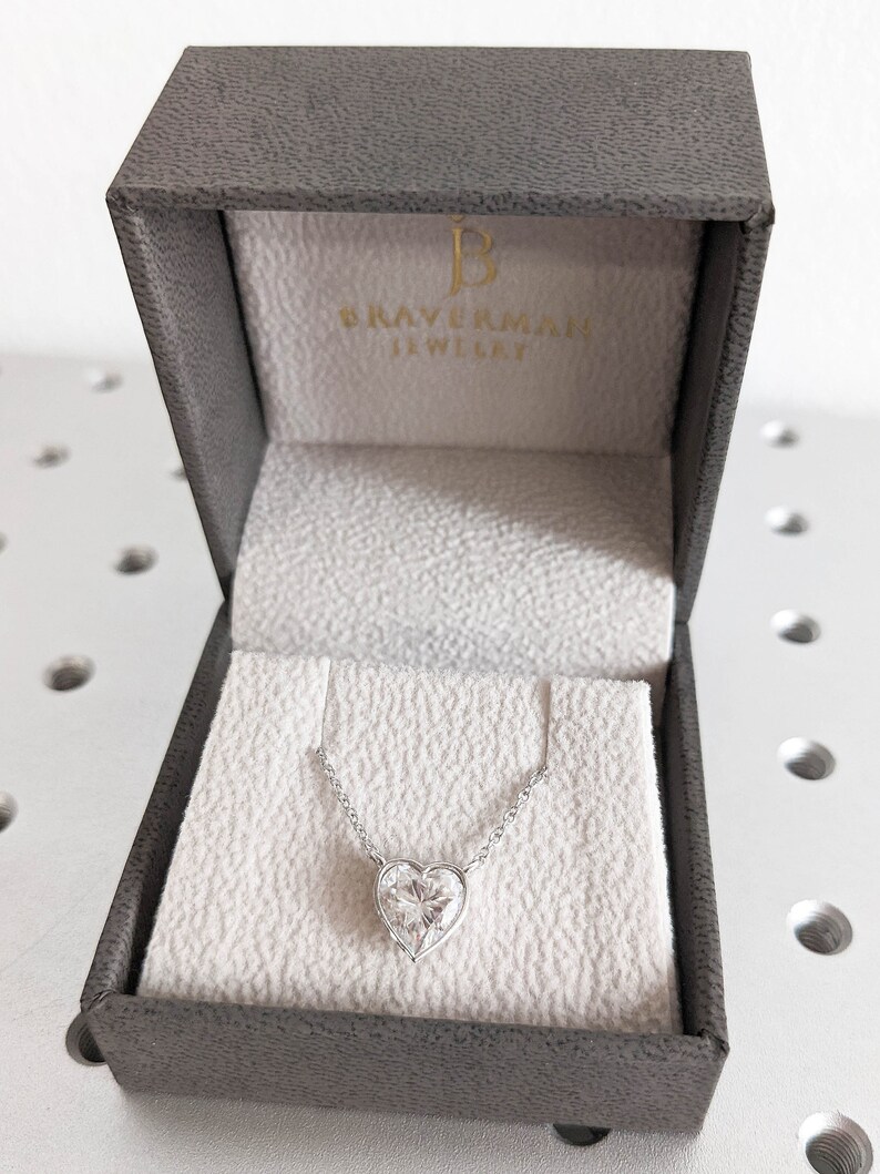Puede incluir: Un collar de plata con un colgante en forma de coraz&oacute;n con un gran diamante transparente en una caja de joyer&iacute;a gris con el texto "BRAVERMAN JEWELLERY" en el interior de la tapa.