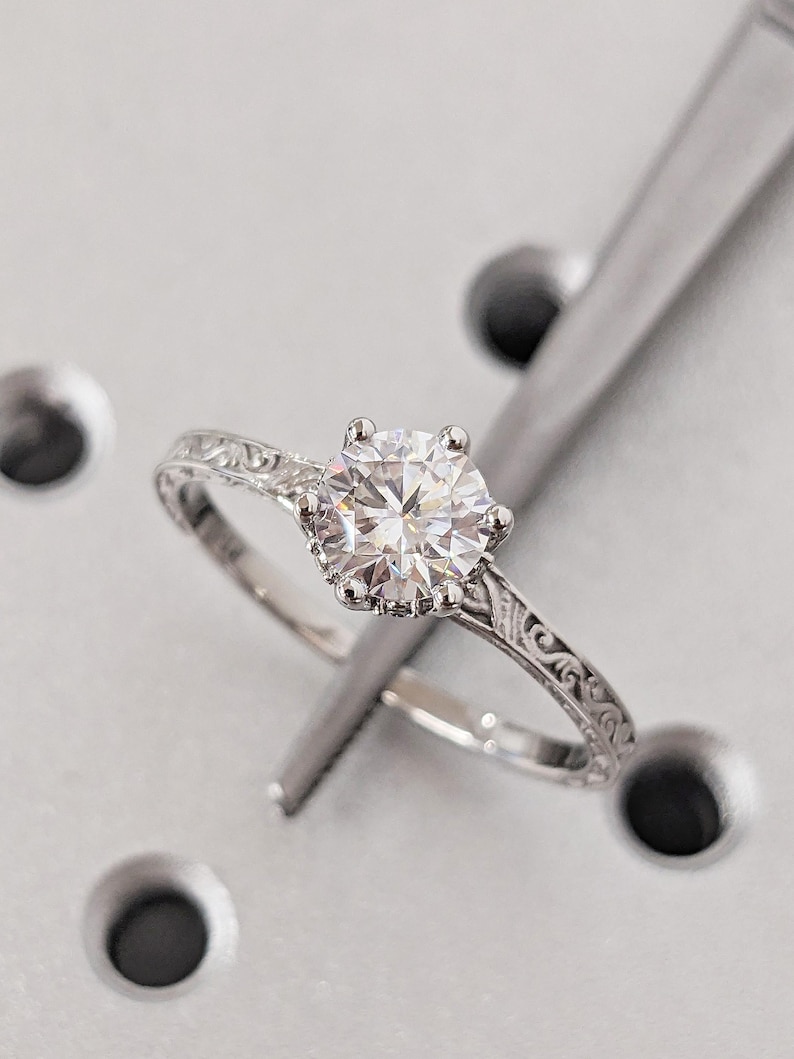 Pu&ograve; includere: Primo piano di un anello di fidanzamento in argento con una grande gemma rotonda e trasparente. L'anello ha una fascia incisa e ornata. L'anello &egrave; posto su uno sfondo grigio chiaro con accenti circolari grigio scuro.