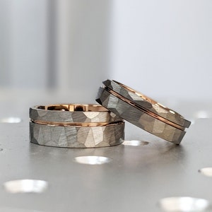 Puede incluir: Tres anillos de boda con un exterior texturizado gris y una banda interior de oro rosa. Los anillos están apilados e inclinados, mostrando el contraste entre los tonos grises y dorados. Los anillos están sobre una superficie reflectante.