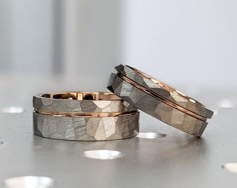 May include: Three wedding rings with a textured, gray exterior and a rose gold interior band. The rings are stacked and angled, showcasing the contrast between the gray and gold tones. The rings are on a reflective surface.