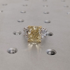 May include: A radiant-cut yellow diamond ring with two baguette-cut side stones. The ring features a platinum band and a gold setting for the center stone. The ring is set on a textured, metallic surface.