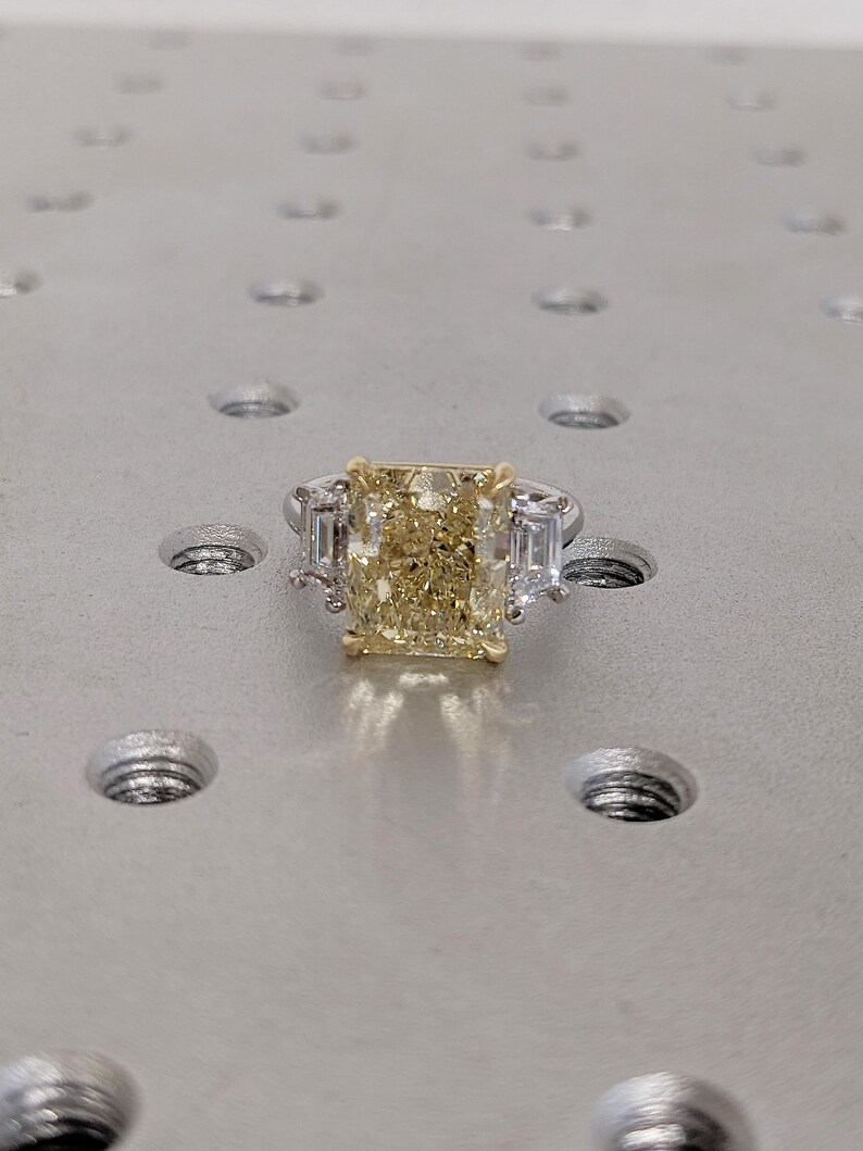 May include: A radiant-cut yellow diamond ring with two baguette-cut side stones. The ring features a platinum band and a gold setting for the center stone. The ring is set on a textured, metallic surface.
