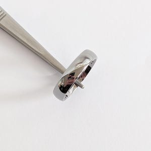 May include: A polished silver ring with a rounded band is held by a silver tool. The ring reflects the surrounding environment. The ring is a simple, elegant design, suitable for everyday wear.