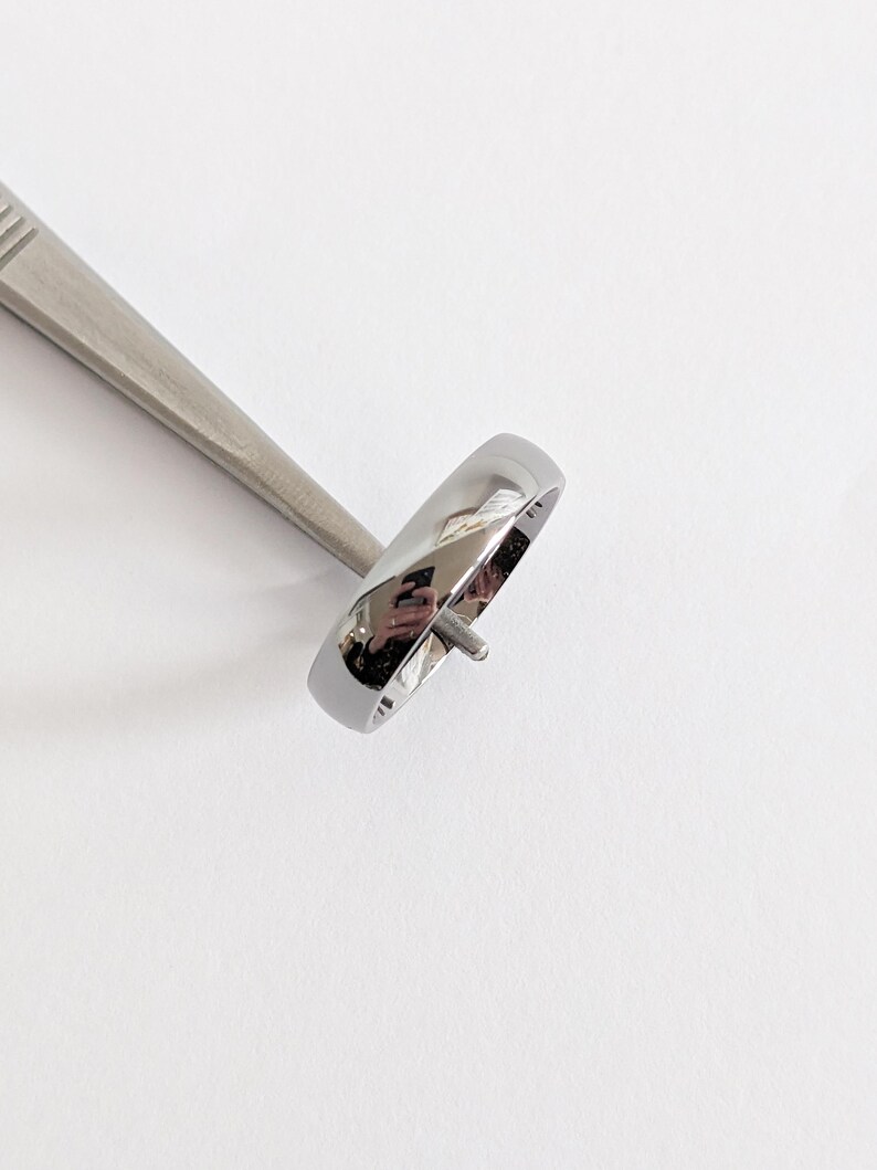 May include: A polished silver ring with a rounded band is held by a silver tool. The ring reflects the surrounding environment. The ring is a simple, elegant design, suitable for everyday wear.