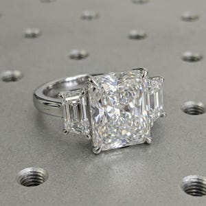 May include: A radiant-cut diamond engagement ring with a platinum band. The ring features a large, rectangular center stone flanked by two smaller emerald-cut diamonds. The diamonds sparkle brightly, showcasing their clarity and cut.
