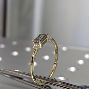 May include: A gold ring with a rectangular grey gemstone and two smaller clear gemstones. The ring is on a silver surface.