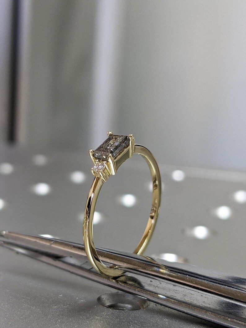 May include: A gold ring with a rectangular grey gemstone and two smaller clear gemstones. The ring is on a silver surface.