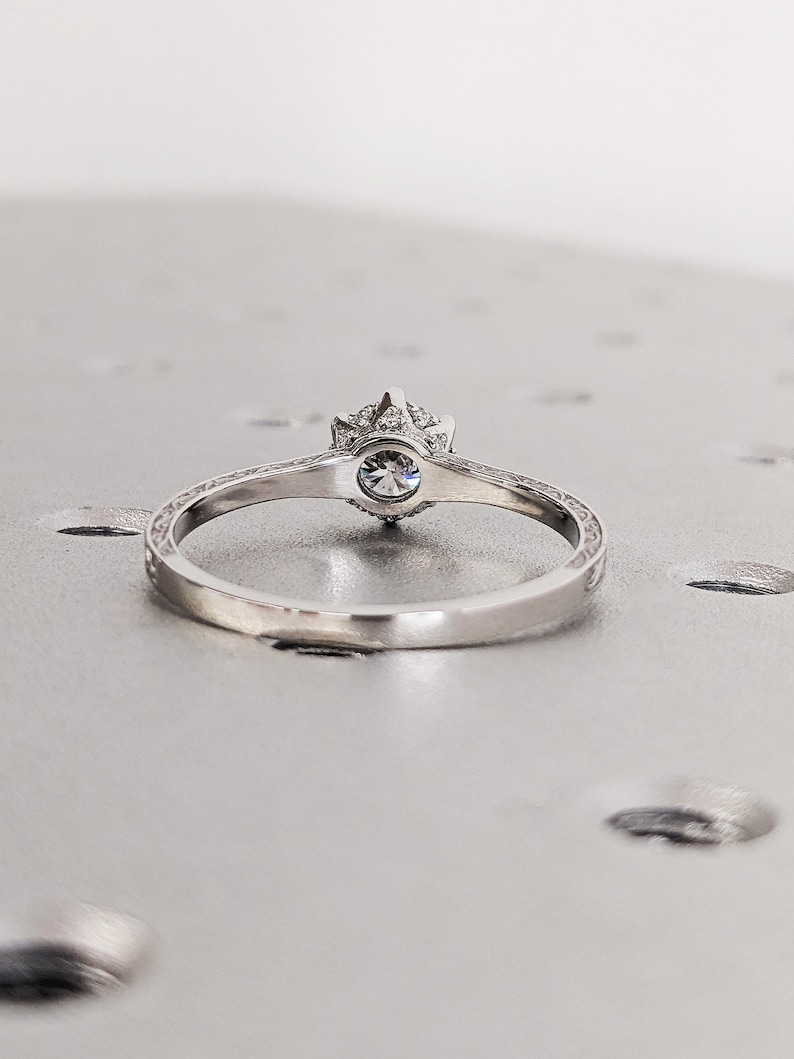 Pu&ograve; includere: Un anello di fidanzamento in argento con una gemma rotonda e trasparente al centro. L'anello presenta una fascia decorativa dettagliata e un alone di pietre pi&ugrave; piccole attorno alla gemma principale. L'anello &egrave; posto su uno sfondo grigio chiaro.