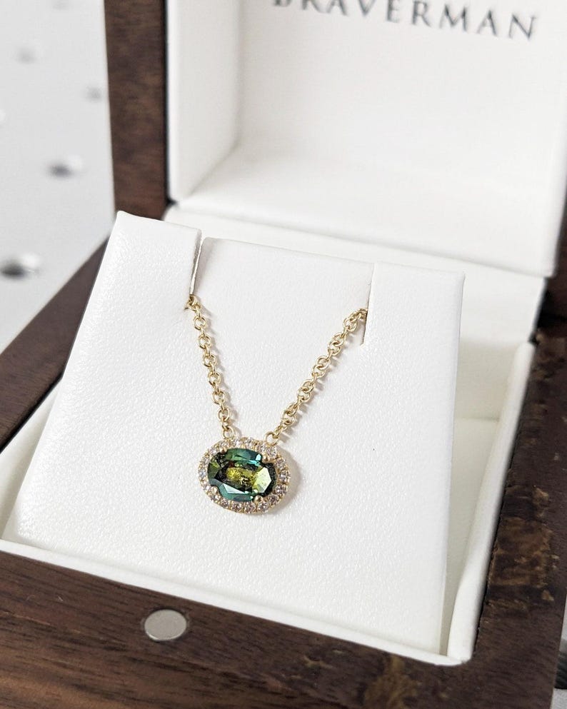 May include: A gold chain necklace with a green oval gemstone surrounded by a halo of smaller diamonds. The necklace is displayed in a wooden box with the name "BRAVERMAN" on the lid.