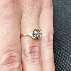 Puede incluir: Un anillo de oro con un gran diamante gris redondo rodeado de dos diamantes triangulares m&aacute;s peque&ntilde;os.