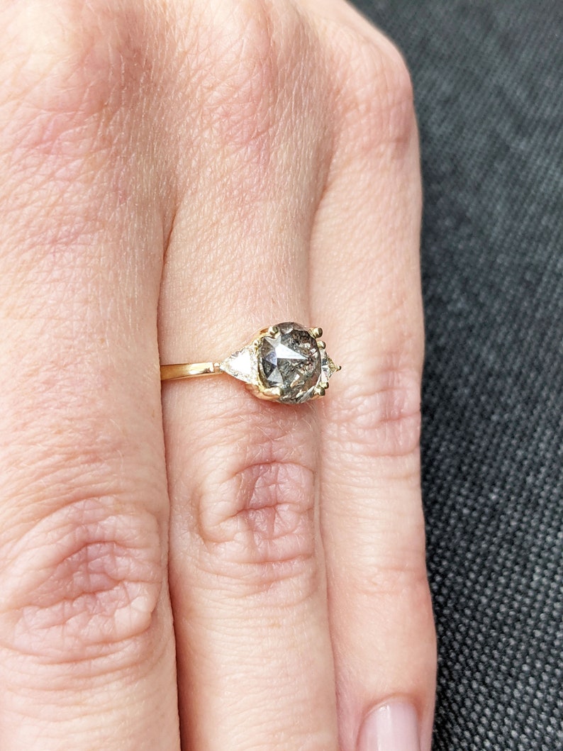 Puede incluir: Un anillo de oro con un gran diamante gris redondo rodeado de dos diamantes triangulares m&aacute;s peque&ntilde;os.