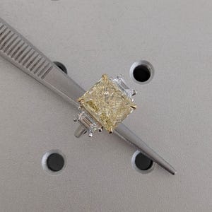 May include: A radiant-cut yellow diamond ring with a gold band and two smaller, clear baguette diamonds on each side. The ring is held by a silver tweezer against a gray background.