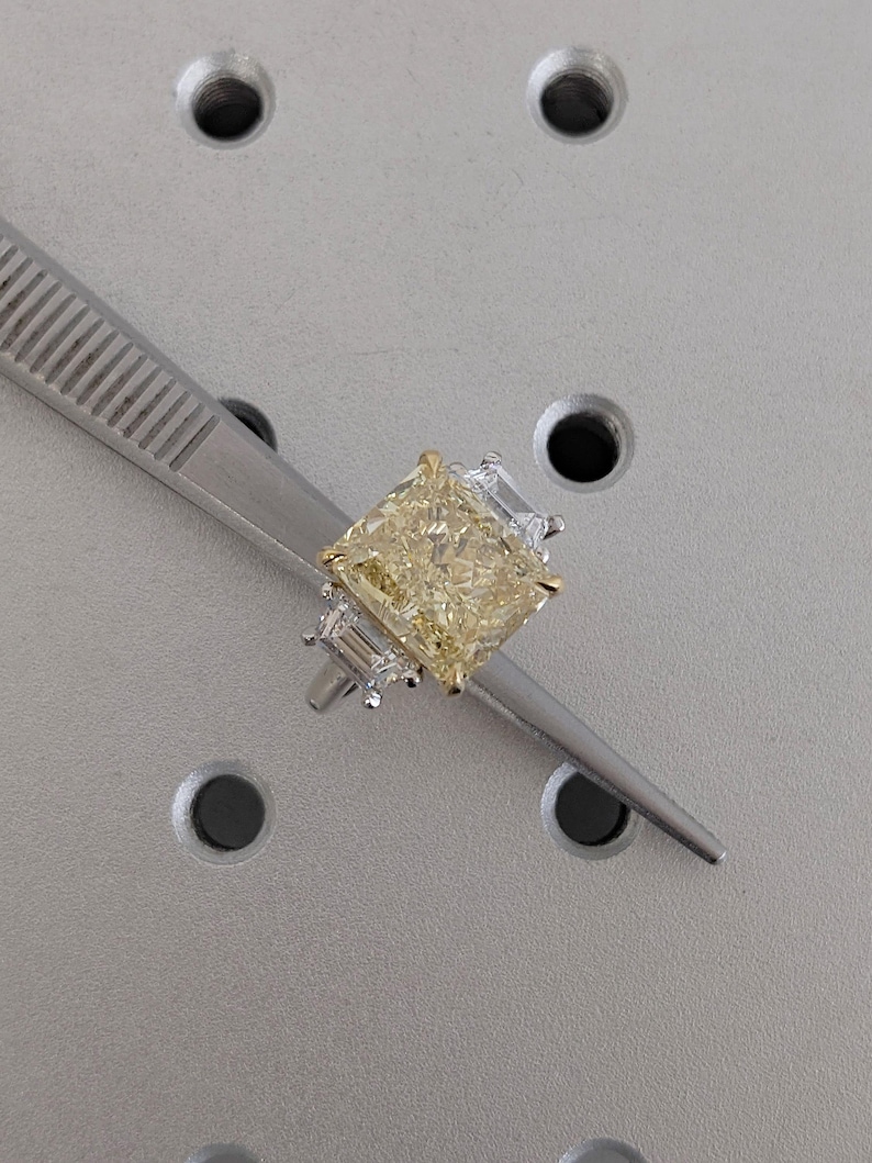 May include: A radiant-cut yellow diamond ring with a gold band and two smaller, clear baguette diamonds on each side. The ring is held by a silver tweezer against a gray background.