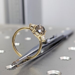 Puede incluir: Un anillo de compromiso de oro con un gran diamante gris redondo y diamantes m&aacute;s peque&ntilde;os en la banda. El anillo est&aacute; sujeto por unas pinzas sobre una superficie met&aacute;lica.