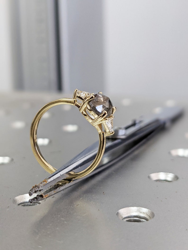 Puede incluir: Un anillo de compromiso de oro con un gran diamante gris redondo y diamantes m&aacute;s peque&ntilde;os en la banda. El anillo est&aacute; sujeto por unas pinzas sobre una superficie met&aacute;lica.