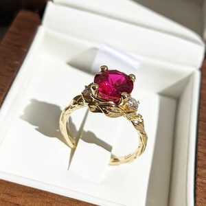 Könnte beinhalten: Ein goldener Ring mit einem großen roten Edelstein und kleineren weißen Edelsteinen, die in einem Blattdesign gefasst sind.