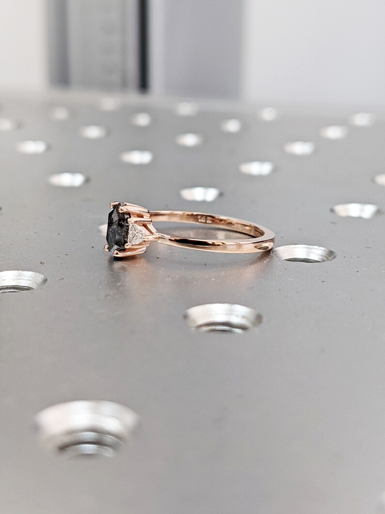 May include: A rose gold engagement ring with a black oval-shaped center stone and two smaller white accent stones.