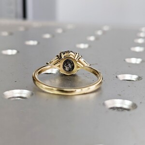Puede incluir: Un anillo de oro con una piedra preciosa redonda grande de color gris rodeada de dos piedras preciosas m&aacute;s peque&ntilde;as en forma de pera.