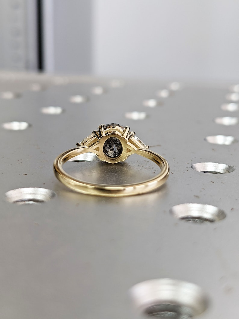Puede incluir: Un anillo de oro con una piedra preciosa redonda grande de color gris rodeada de dos piedras preciosas m&aacute;s peque&ntilde;as en forma de pera.