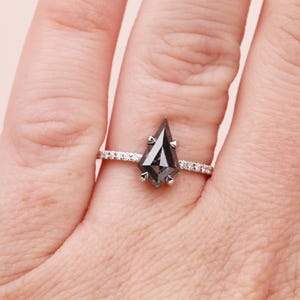 Puede incluir: Un anillo de plata con una gema gris oscura en forma de cometa. La banda est&aacute; adornada con peque&ntilde;as piedras claras. El anillo se muestra en un dedo, destacando su dise&ntilde;o y brillo.
