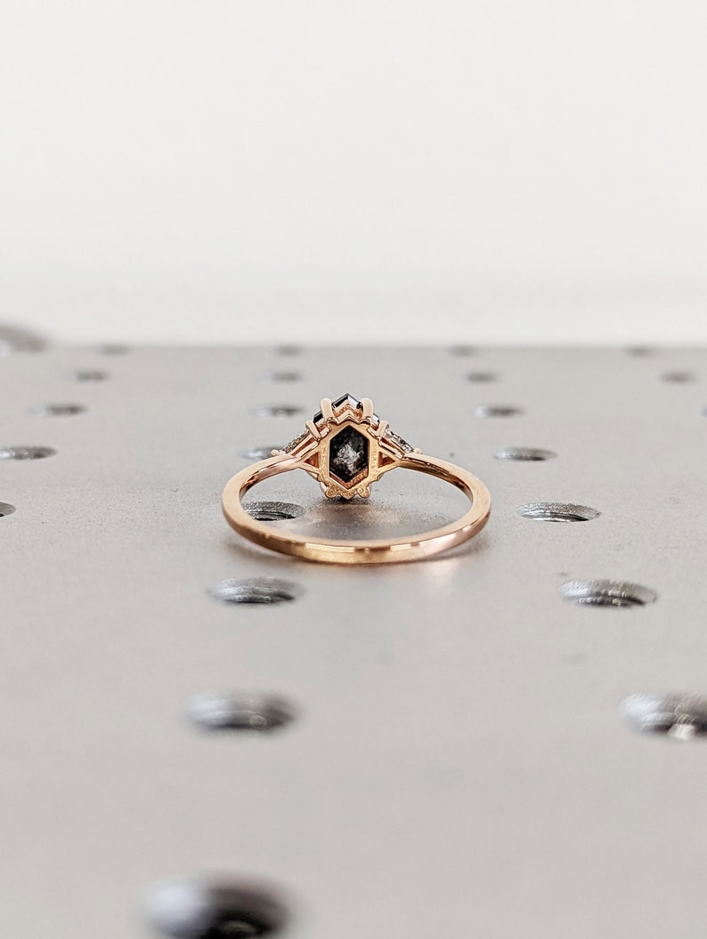 May include: A rose gold ring with a hexagonal shaped black diamond center stone and smaller diamonds on the band.