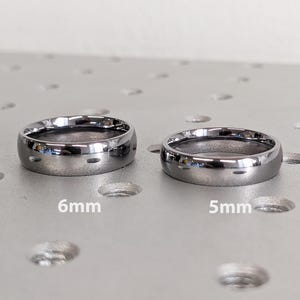 May include: Two silver-colored rings, one 6mm and the other 5mm wide, are displayed. The rings have a polished finish and a rounded shape. The image is taken on a textured, light-colored surface.