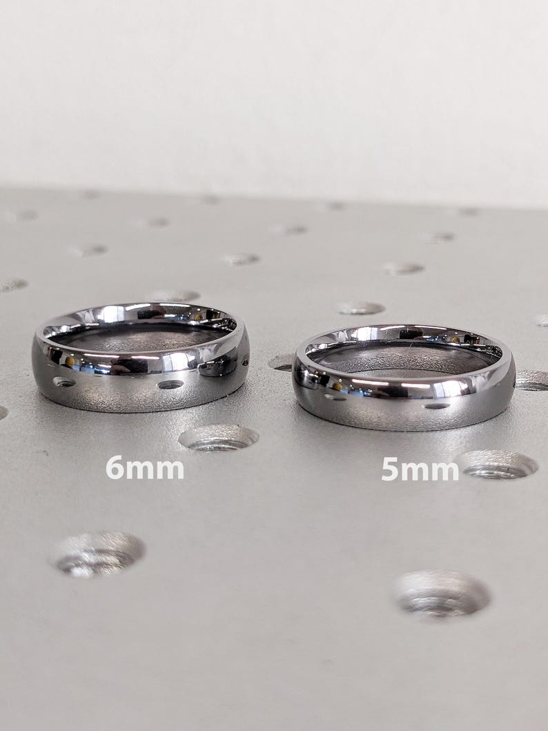 May include: Two silver-colored rings, one 6mm and the other 5mm wide, are displayed. The rings have a polished finish and a rounded shape. The image is taken on a textured, light-colored surface.