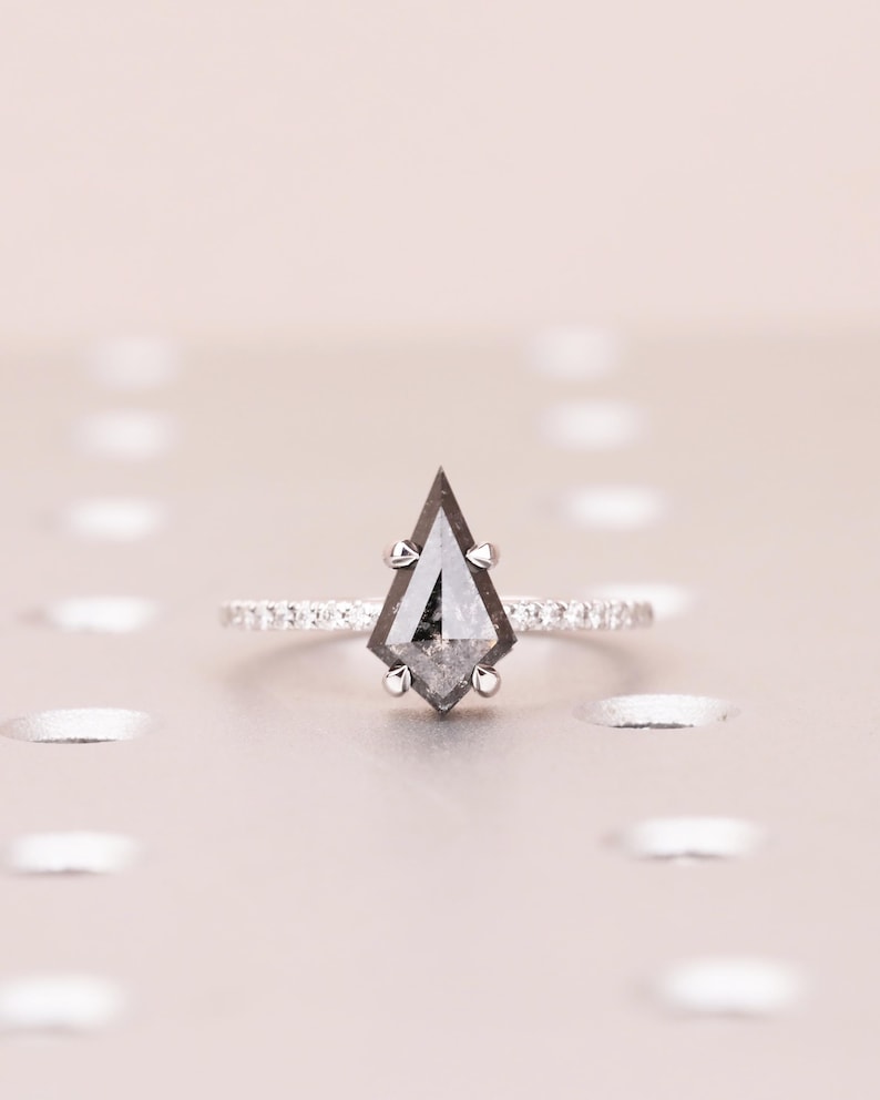 Puede incluir: Un anillo de plata con un diamante gris oscuro en forma de cometa. La banda est&aacute; adornada con peque&ntilde;as piedras brillantes. El dise&ntilde;o del anillo se muestra sobre un fondo suave y neutro.