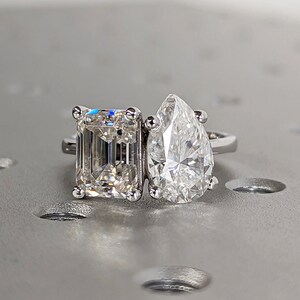 May include: A silver ring featuring two large diamonds. The diamond on the left is a square emerald cut, and the diamond on the right is a pear-shaped diamond.