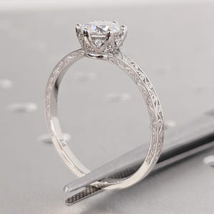 Pu&ograve; includere: Primo piano di un anello di fidanzamento in platino con una grande gemma rotonda e trasparente. L'anello presenta un intricato design inciso sulla fascia ed &egrave; tenuto da una pinzetta.