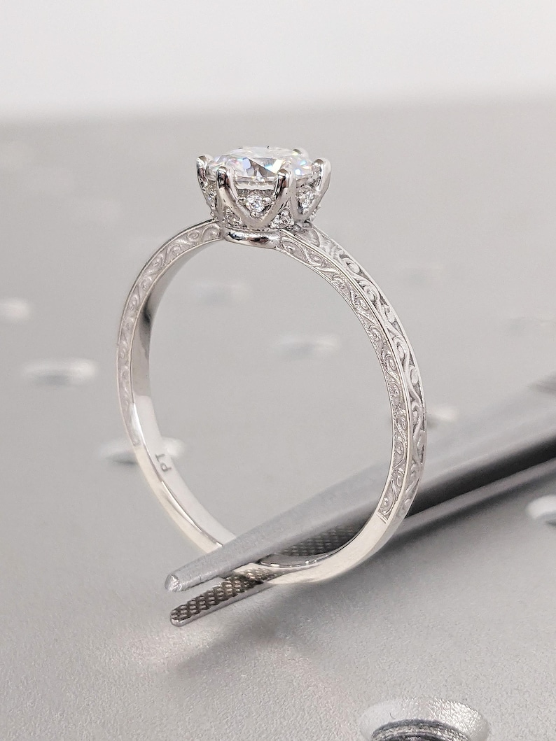 Pu&ograve; includere: Primo piano di un anello di fidanzamento in platino con una grande gemma rotonda e trasparente. L'anello presenta un intricato design inciso sulla fascia ed &egrave; tenuto da una pinzetta.