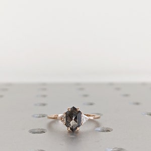 May include: A rose gold engagement ring with a hexagonal gray diamond and two smaller triangular diamonds on either side.