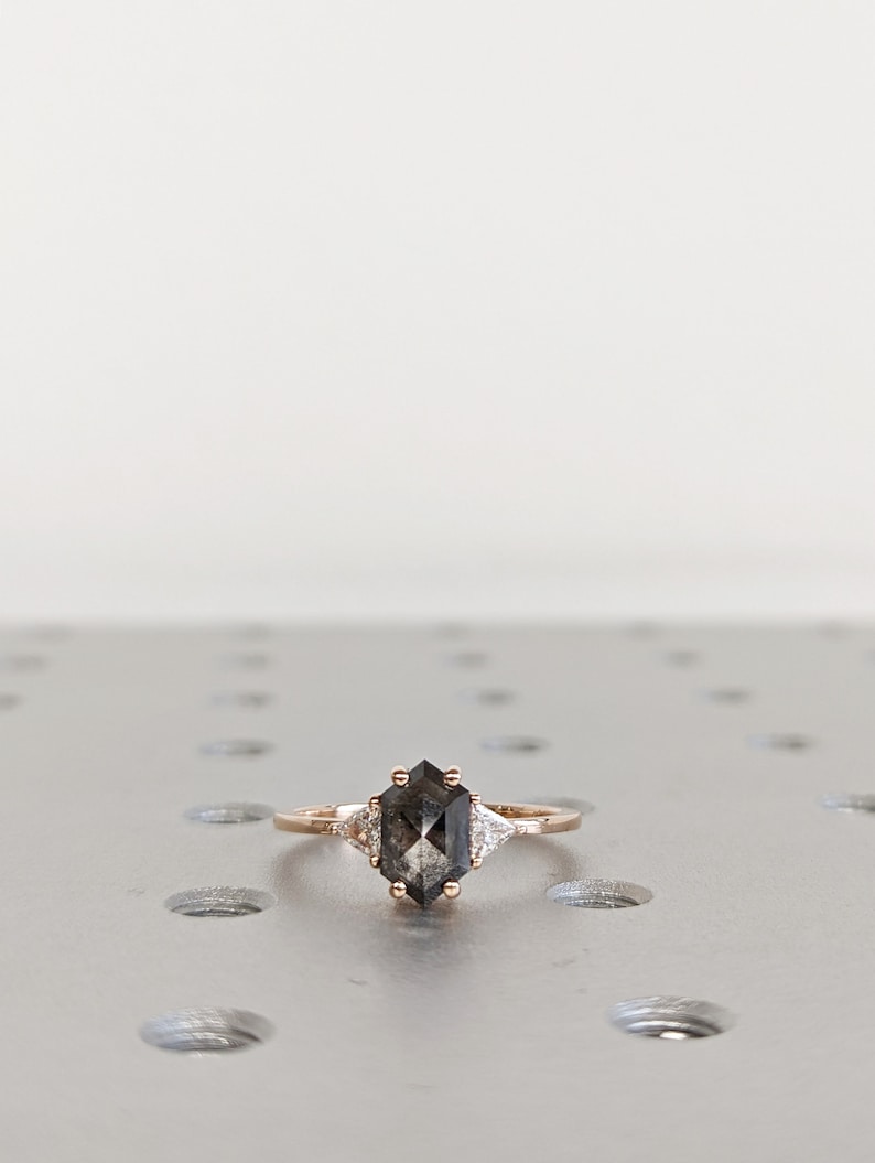 May include: A rose gold engagement ring with a hexagonal gray diamond and two smaller triangular diamonds on either side.