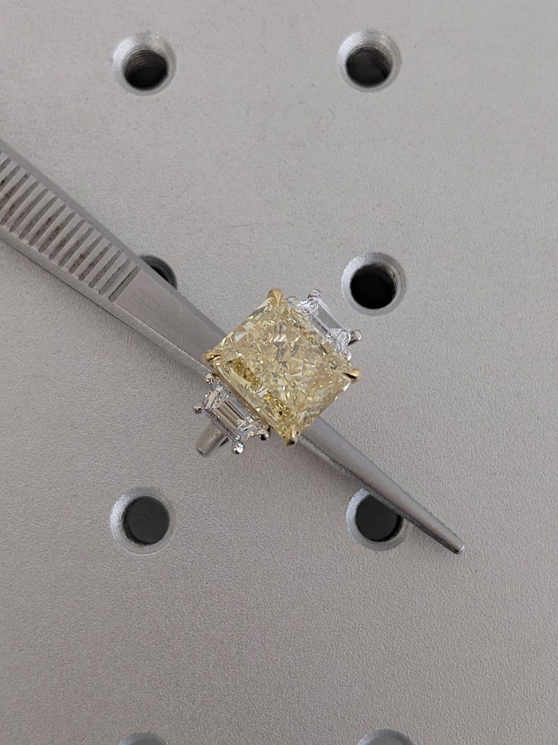 May include: A yellow radiant-cut diamond ring with a gold band and two smaller, clear side stones. The ring is held by a silver tweezer against a grey background. The diamond has a square shape with a brilliant cut.