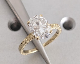 May include: A gold engagement ring with an oval-cut diamond. The ring features an engraved band with a floral pattern. The diamond is held in place by four prongs. The ring is set against a light gray background.
