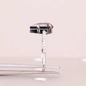 Puede incluir: Un anillo de plata con una piedra preciosa gris oscura en forma de pera. La banda est&aacute; adornada con peque&ntilde;as piedras transparentes. El anillo est&aacute; sujeto por unas pinzas plateadas, sobre un fondo rosa claro.