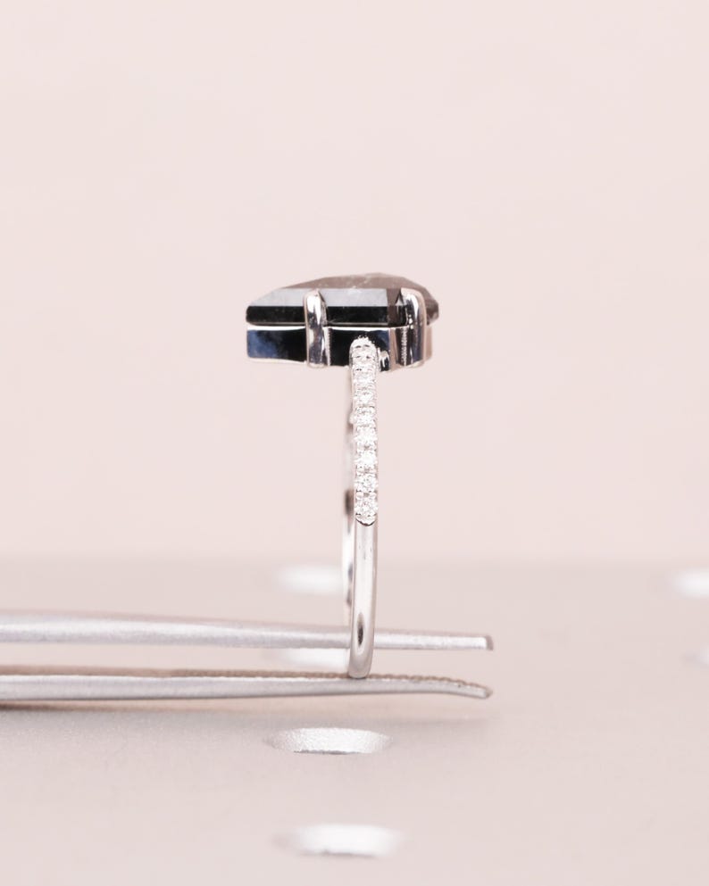Puede incluir: Un anillo de plata con una piedra preciosa gris oscura en forma de pera. La banda est&aacute; adornada con peque&ntilde;as piedras transparentes. El anillo est&aacute; sujeto por unas pinzas plateadas, sobre un fondo rosa claro.