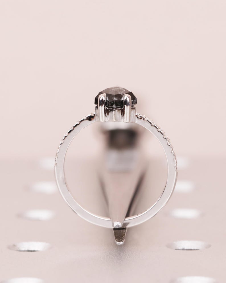 Puede incluir: Un anillo plateado con una gema grande de color gris oscuro en el centro. La banda est&aacute; adornada con peque&ntilde;as piedras claras. El anillo se presenta sobre un fondo rosa claro, creando un contraste.