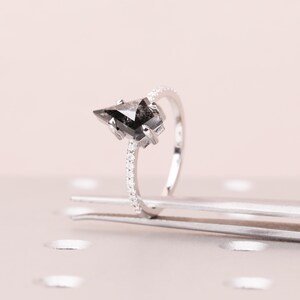Puede incluir: Un anillo de plata con una piedra preciosa negra triangular. La banda est&aacute; adornada con peque&ntilde;as piedras claras. El anillo se muestra sobre una superficie rosa claro, resaltando su elegante dise&ntilde;o. Una joya contempor&aacute;nea.