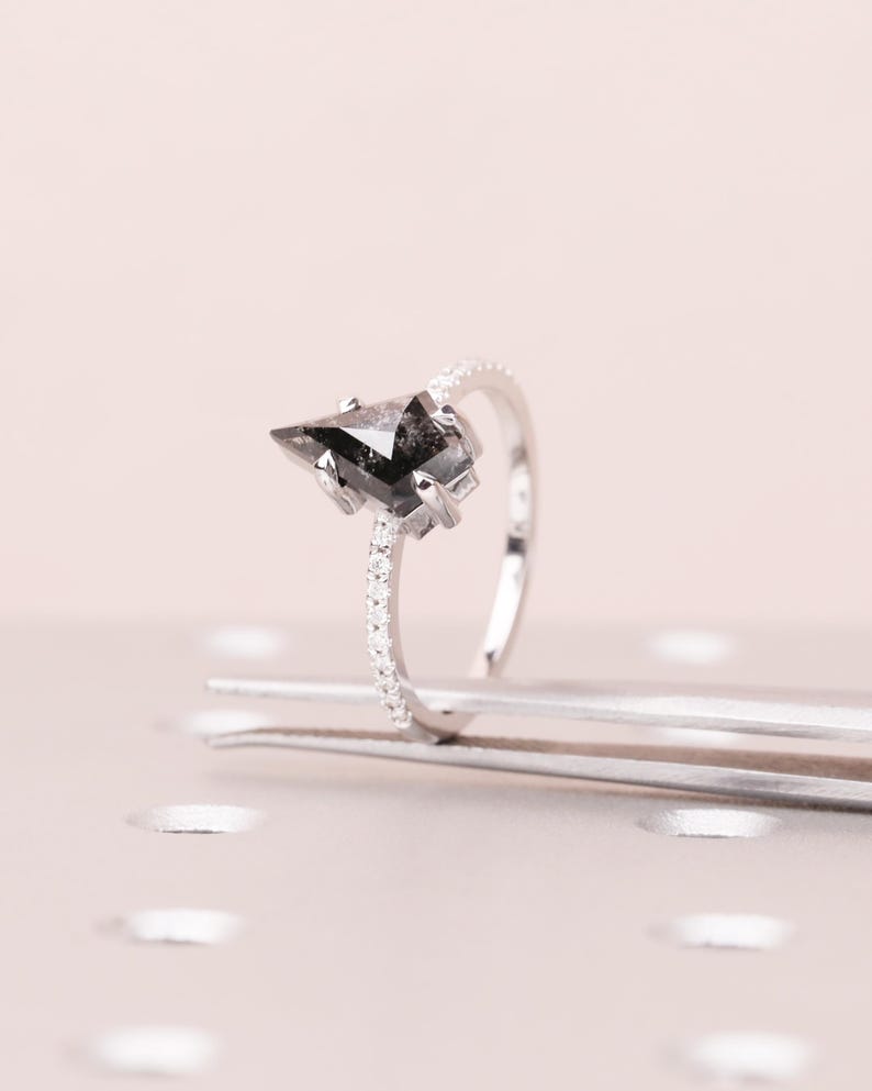 Puede incluir: Un anillo de plata con una piedra preciosa negra triangular. La banda est&aacute; adornada con peque&ntilde;as piedras claras. El anillo se muestra sobre una superficie rosa claro, resaltando su elegante dise&ntilde;o. Una joya contempor&aacute;nea.
