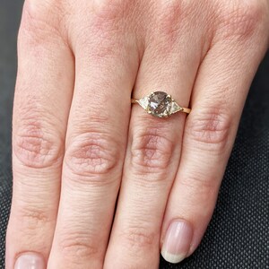 Puede incluir: Un anillo de oro con un diamante redondo gris en el centro y dos diamantes triangulares m&aacute;s peque&ntilde;os a cada lado.