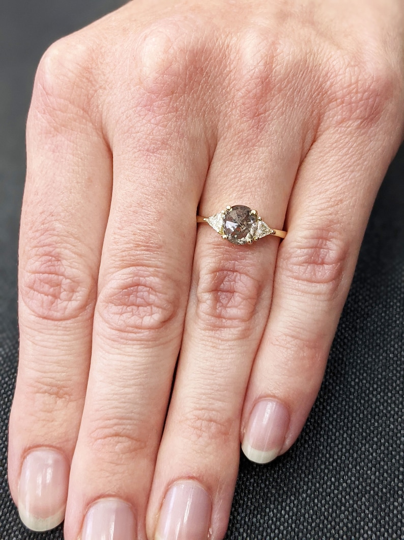 Puede incluir: Un anillo de oro con un diamante redondo gris en el centro y dos diamantes triangulares m&aacute;s peque&ntilde;os a cada lado.