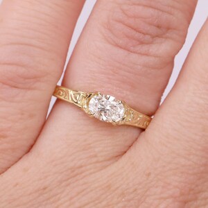 Puede incluir: Un anillo de oro con una piedra preciosa transparente de corte ovalado engastada en el centro. La banda está hecha de oro amarillo y presenta un intrincado patrón de volutas grabadas que se extiende por los lados del anillo, rodeando la piedra.