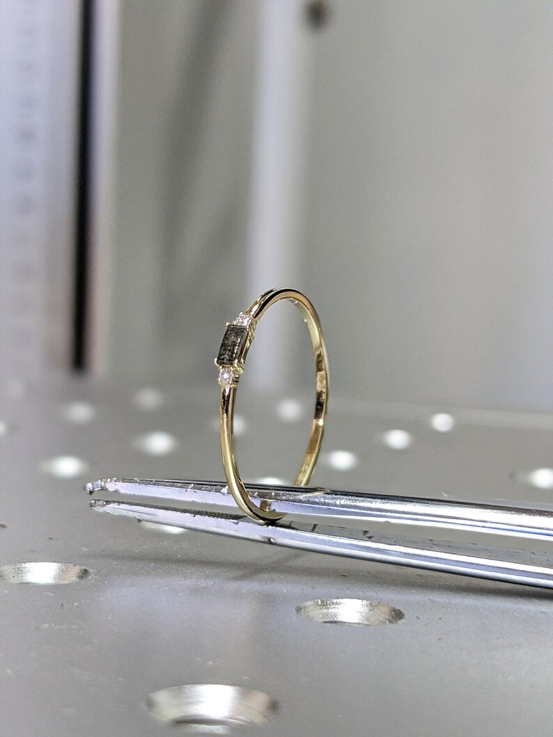May include: A delicate gold band ring with a small rectangular black diamond and two smaller diamonds on either side.