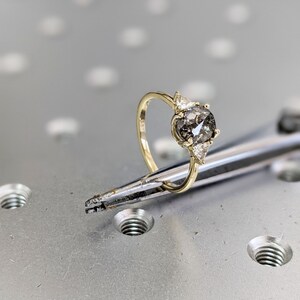 Puede incluir: Un anillo de compromiso de oro con un gran diamante gris redondo y dos diamantes triangulares m&aacute;s peque&ntilde;os a cada lado.