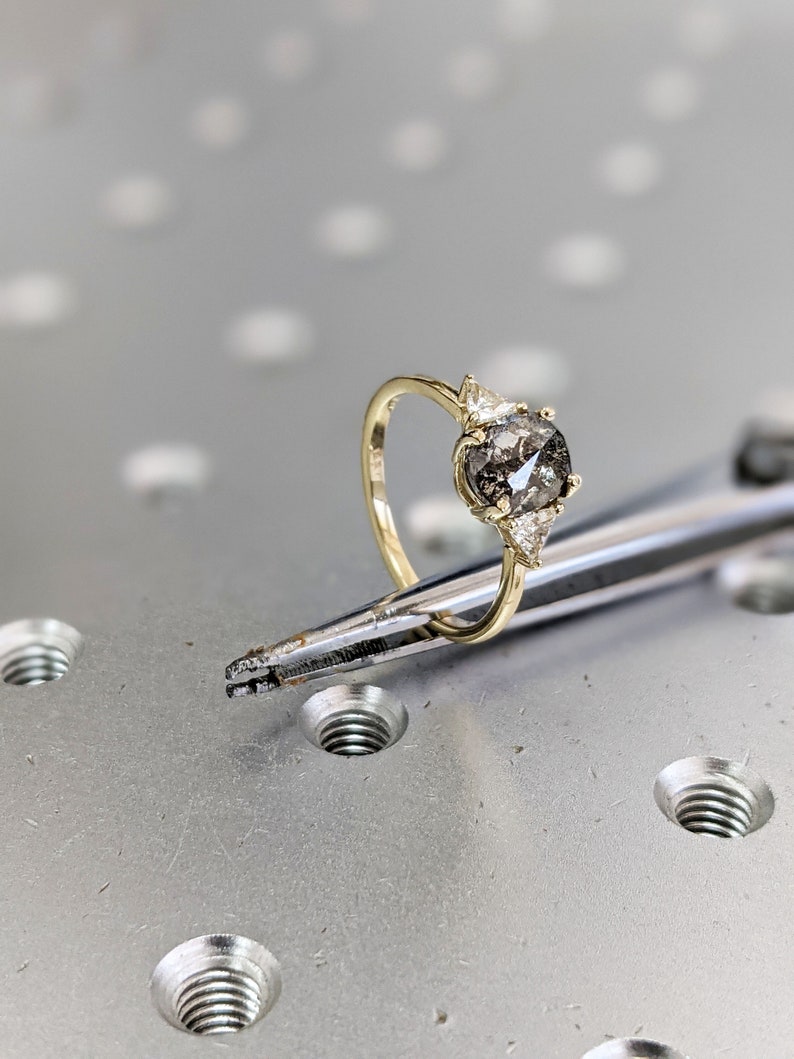 Puede incluir: Un anillo de compromiso de oro con un gran diamante gris redondo y dos diamantes triangulares m&aacute;s peque&ntilde;os a cada lado.